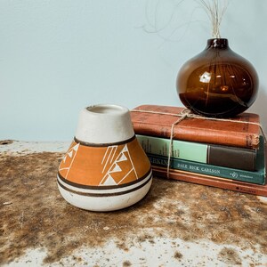 Può includere: Un vaso di ceramica con la parte superiore bianca e un design geometrico arancione. Un piccolo vaso di vetro marrone con erba secca è posto sopra una pila di libri d'epoca. I libri sono legati con spago.