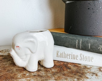Puede incluir: Una pequeña figura de elefante de cerámica blanca con detalles rojos en las orejas. El elefante está sobre una superficie metálica oxidada y desgastada. En el fondo hay libros y un jarrón oscuro.