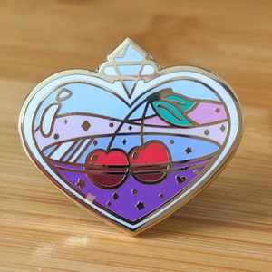 Op de afbeelding: Gouden hartvormige emaille pin met een blauw, paars en roze ontwerp. De pin heeft twee rode kersen in een glazen fles met een gouden deksel.