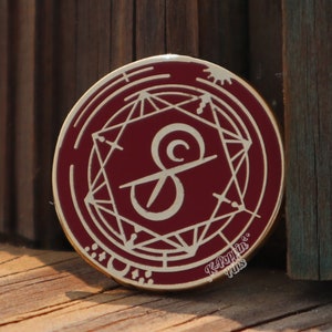 May include: A gold-colored enamel pin with a red background. The pin features a stylized letter "S" inside a circle with geometric designs. The text "Jay Build Things" is printed on the pin.
