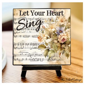 May include: A square decorative sign with the words "Let Your Heart Sing" over a vintage sheet music background. The sign features a floral arrangement with roses and other flowers in soft colors, displayed on a small black easel.