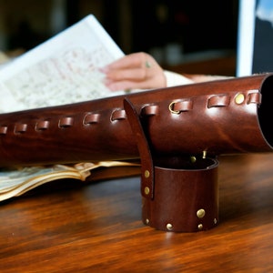 May include: A dark brown leather telescope case with brass detailing. The cylindrical case features a handle and a separate, smaller cylindrical piece. It rests on a wooden surface.