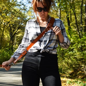 May include: A woman wearing a black and white checked shirt with a brown leather camera strap. She is wearing black jeans and sunglasses.
