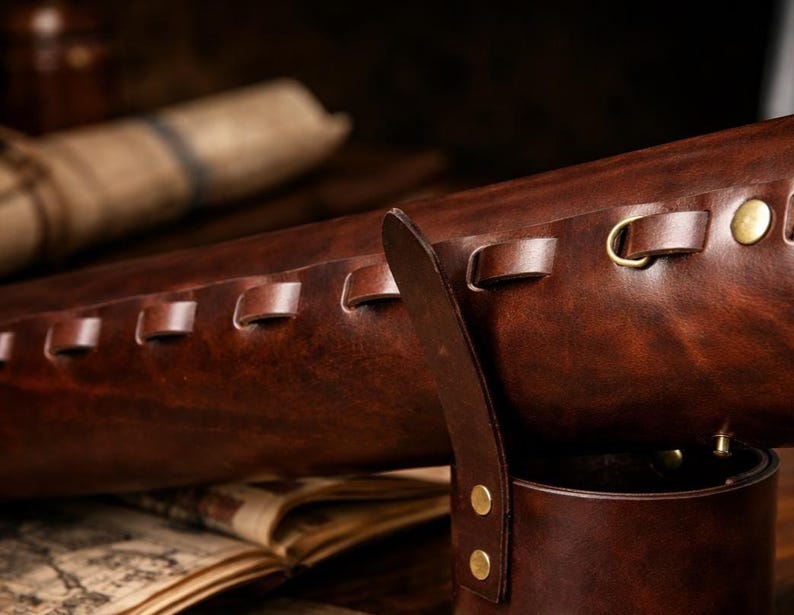 May include: A close-up of a brown leather quiver with brass accents. The quiver has a cylindrical shape with a strap and is decorated with stitched details and brass rivets. A rolled-up parchment is visible in the background.