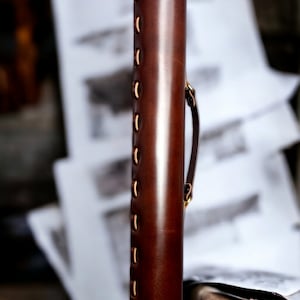 May include: A cylindrical, dark brown leather case with brass rivets and stitching. The case has a handle and a flap closure. The background is blurred, but appears to be a wooden surface and some papers.