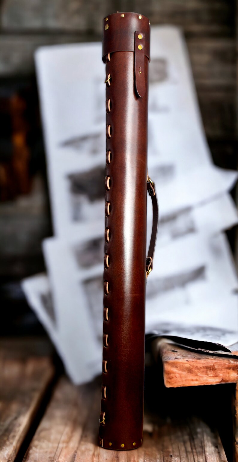 May include: A cylindrical, dark brown leather case with brass rivets and stitching. The case has a handle and a flap closure. The background is blurred, but appears to be a wooden surface and some papers.