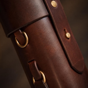 May include: Close-up of a brown leather case with brass hardware. The case has a rolled top, a strap with brass rivets, and gold-coloured metal rings. The leather has a rich, textured appearance.