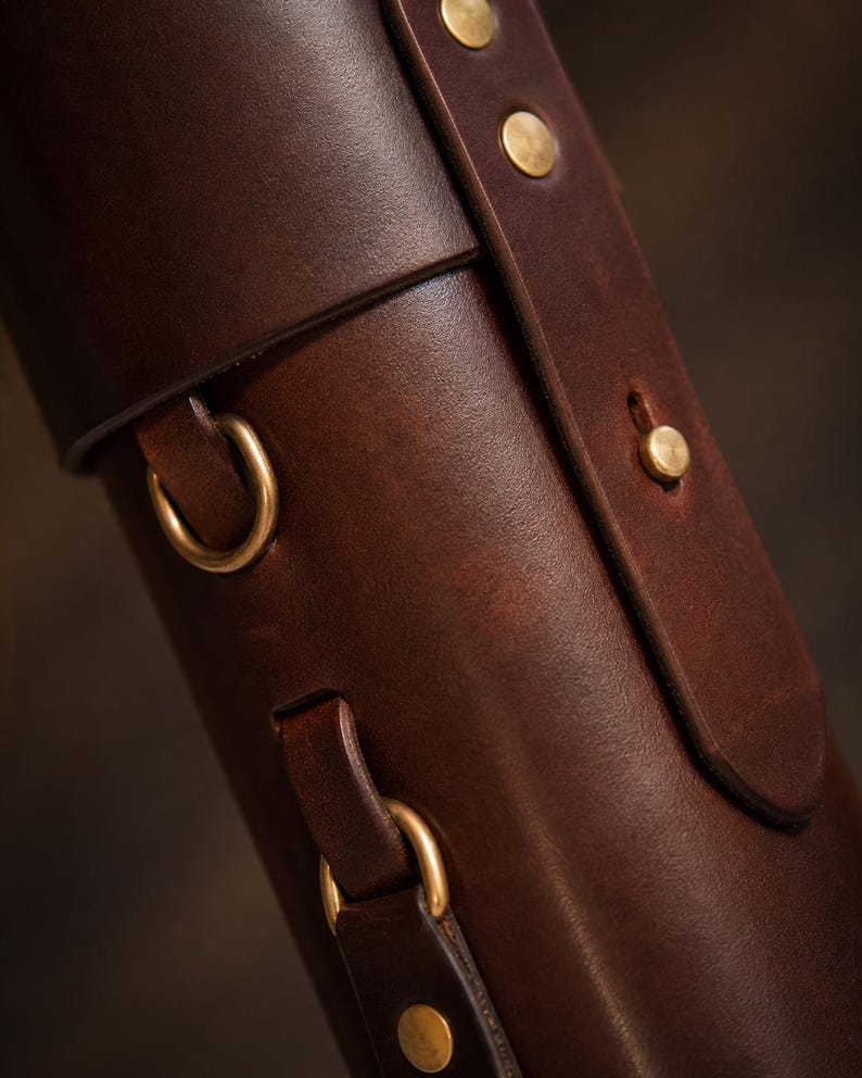 May include: Close-up of a brown leather case with brass hardware. The case has a rolled top, a strap with brass rivets, and gold-coloured metal rings. The leather has a rich, textured appearance.