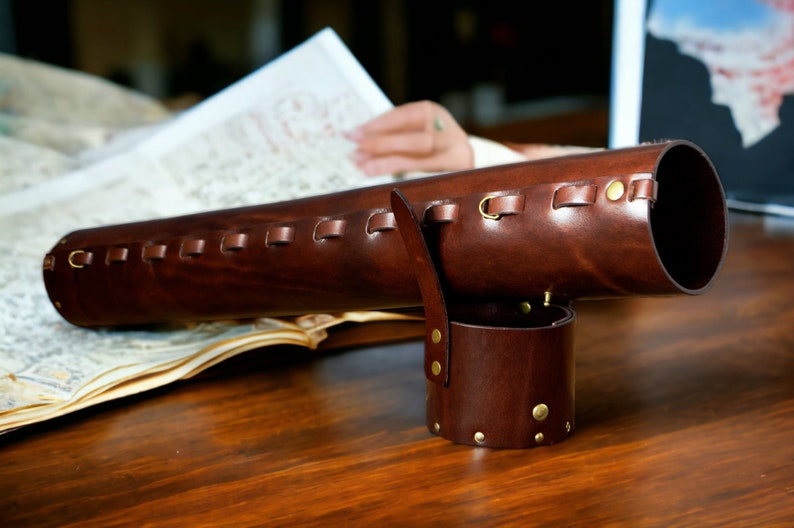 May include: A dark brown leather telescope case with brass detailing. The cylindrical case features a handle and a separate, smaller cylindrical piece. It rests on a wooden surface.