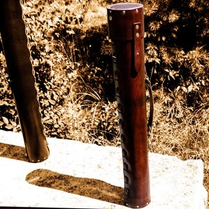 May include: A cylindrical, brown leather quiver with a handle and decorative stitching. The quiver is standing upright on a white surface, with a natural background of foliage.