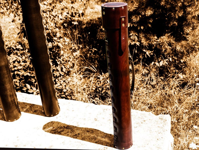 May include: A cylindrical, brown leather quiver with a handle and decorative stitching. The quiver is standing upright on a white surface, with a natural background of foliage.