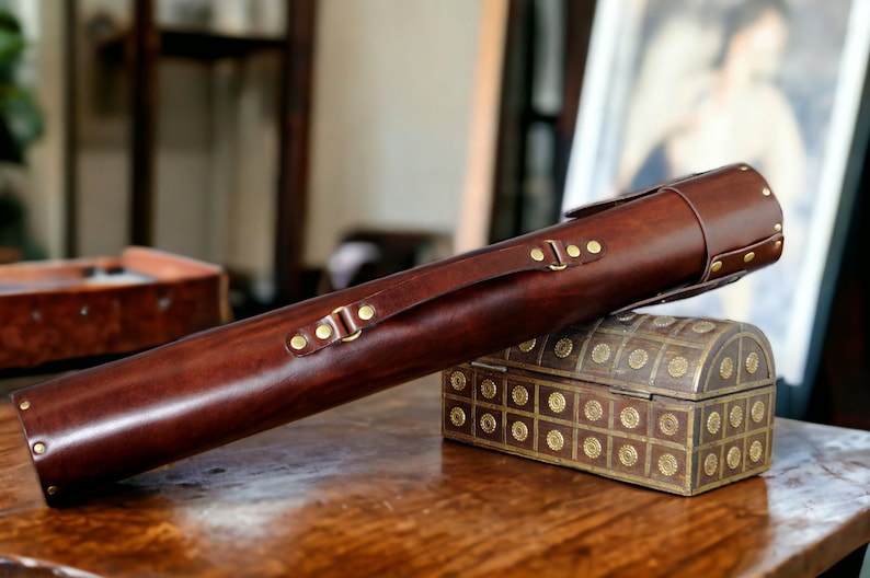 May include: A dark brown leather case with brass fittings and a handle. The cylindrical case is placed on a small wooden chest with brass detailing. The case is likely designed to hold a long, slender item, such as a cue.