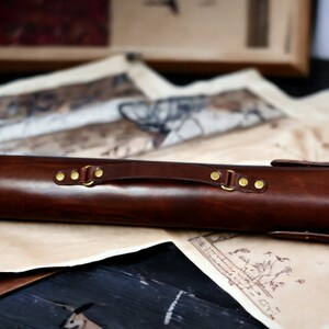 May include: A dark brown leather case with brass fittings, including rivets and a handle. The cylindrical case is designed to hold a long, slender object. The case rests on a surface with vintage-style maps.