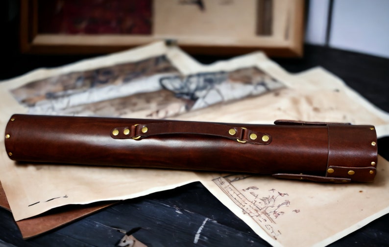May include: A dark brown leather case with brass fittings, including rivets and a handle. The cylindrical case is designed to hold a long, slender object. The case rests on a surface with vintage-style maps.