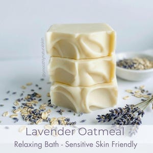Puede incluir: Tres barras de jabón apiladas, de color claro, con un diseño en espiral. Las barras están etiquetadas como "Lavender Oatmeal" y "Relaxing Bath - Sensitive Skin Friendly". Lavanda seca y avena están esparcidas alrededor del jabón, con un pequeño cuenco de avena en el fondo.