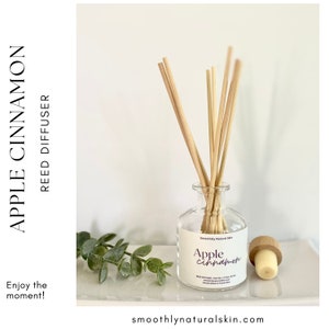 May include: A clear glass reed diffuser with bamboo reeds, a wooden stopper, and a label that reads "Apple Cinnamon." The diffuser sits on a white tray with a sprig of greenery. The text "Enjoy the moment!" is also visible.