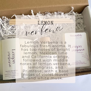 May include: A box with a white and brown paper filler. The box contains three products: a candle, a soy wax melt, and a diffuser. The candle is clear glass with a white label that says 'Lemon Verbena' and 'Smoothly Natural Skin'. The soy wax melt is a small white container with a white label that says 'Lemon Verbena' and 'Smoothly Natural Skin'. The diffuser is a glass bottle with a wooden lid and a white label that says 'Lemon Verbena'. The text on the label reads: 'Lemon Verbena is a fabulous fresh aroma. It has top notes of bright lemon, Mexican lime and California orange, followed with middle notes of lemon verbena, lemongrass, and Primrose; with a base notes of violet leaves and white musk.'