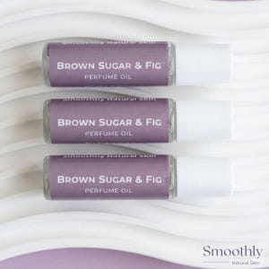 May include: Three cylindrical glass perfume oil bottles with white caps, each featuring a purple label with the text "smoothly Natural Skin" and "BROWN SUGAR & FIG PERFUME OIL" printed in white, arranged horizontally in a parallel stack.
