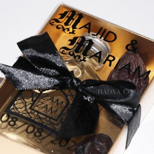 May include: A clear box with a gold background contains a small bottle of perfume, two dates, and a black ribbon. The box has the names "Majid & Mariam" and the date "08/08/202" printed on it. The bottle has a black label with the words "Zam Zam" printed on it. The box also has the words "Hadya Co" printed on it.