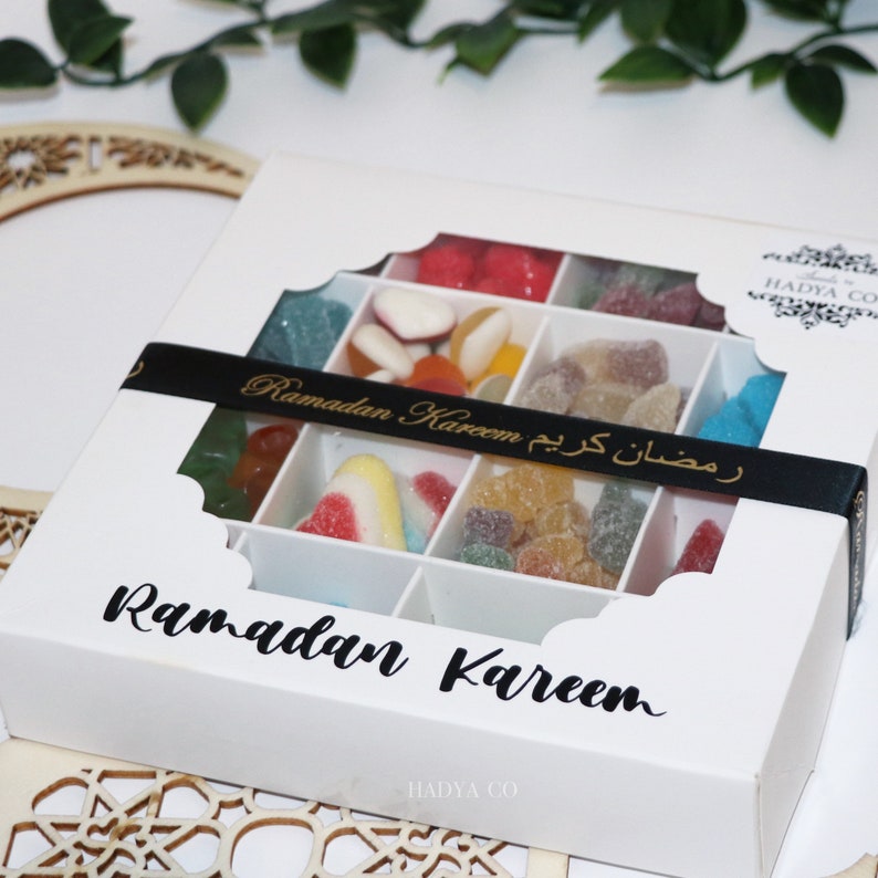 Puede incluir: Caja blanca con tapa calada llena de caramelos coloridos. La caja est&aacute; decorada con las palabras "Ramadan Kareem" en ingl&eacute;s y &aacute;rabe. Una cinta negra con texto dorado envuelve la caja.