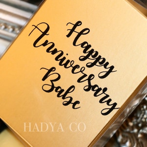May include: Gold gift box with a black inscription that reads "Happy Anniversary Babe" and the text "HADYA CO" at the bottom.