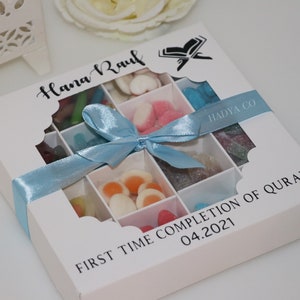 Puede incluir: Una caja de regalo blanca con una cinta azul claro, llena de caramelos de colores variados. La caja tiene un dise&ntilde;o con ventanas y el texto "Hana Raul" y "First Time Completion of Quran 04.2021".