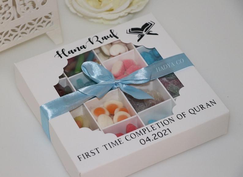 Puede incluir: Una caja de regalo blanca con una cinta azul claro, llena de caramelos de colores variados. La caja tiene un dise&ntilde;o con ventanas y el texto "Hana Raul" y "First Time Completion of Quran 04.2021".