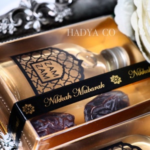 May include: A gold gift box with a clear lid containing a small bottle of Zam Zam water, two dates, and a black ribbon with the text "Nikkah Mubarak" in gold lettering. The box is sitting on a silver tray with a white rose in the background.