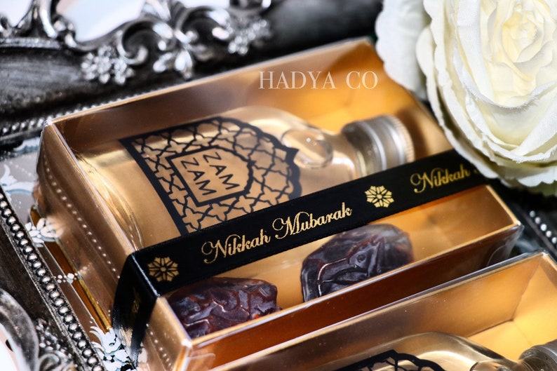 May include: A gold gift box with a clear lid containing a small bottle of Zam Zam water, two dates, and a black ribbon with the text "Nikkah Mubarak" in gold lettering. The box is sitting on a silver tray with a white rose in the background.