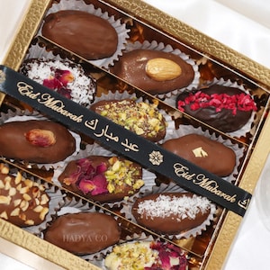 May include: A gold box filled with chocolate-covered dates. The dates are decorated with various toppings, including nuts, coconut flakes, and dried fruit. The box is tied with a black ribbon that says "Eid Mubarak".