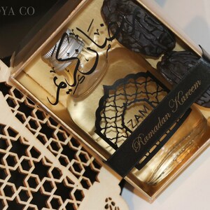 May include: A gold gift box with a clear lid containing a small bottle of perfume, dates, and a black ribbon with the text "Ramadan Kareem".