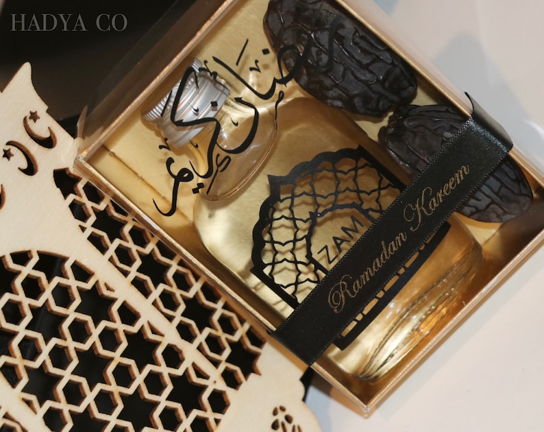 May include: A gold gift box with a clear lid containing a small bottle of perfume, dates, and a black ribbon with the text "Ramadan Kareem".