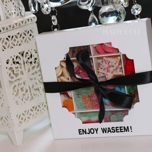 Puede incluir: Una caja de regalo blanca con una ventana decorativa, llena de caramelos variados y atada con una cinta negra. La caja contiene dulces de varias formas y colores. El texto "ENJOY WASEEM!" est&aacute; impreso en la caja.