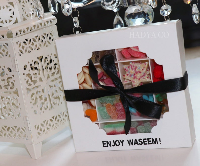 Puede incluir: Una caja de regalo blanca con una ventana decorativa, llena de caramelos variados y atada con una cinta negra. La caja contiene dulces de varias formas y colores. El texto "ENJOY WASEEM!" est&aacute; impreso en la caja.