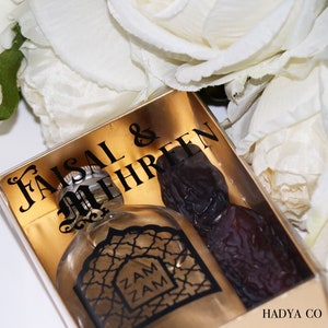 May include: A clear glass bottle with a gold label that says "Zam Zam" inside a clear gift box with a gold background. The box also has a black label that says "Faisal & Mehreen" and a brown date fruit. The box has a white label on the bottom that says "Hadya Co."