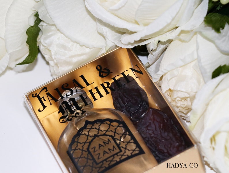 May include: A clear glass bottle with a gold label that says "Zam Zam" inside a clear gift box with a gold background. The box also has a black label that says "Faisal & Mehreen" and a brown date fruit. The box has a white label on the bottom that says "Hadya Co."