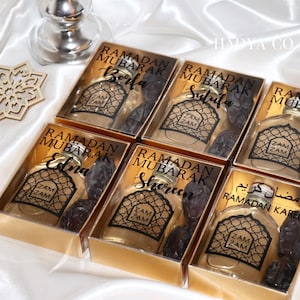 May include: Six gold gift boxes with clear lids containing small bottles of Zam Zam water and dates. Each box has a label with the text "Ramadan Mubarak" and a name, such as "Borfa", "Sutufa", "Edna", "Shereen", and "Ramadan Kareem".