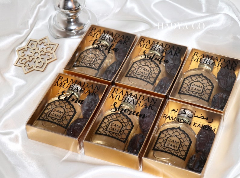 May include: Six gold gift boxes with clear lids containing small bottles of Zam Zam water and dates. Each box has a label with the text "Ramadan Mubarak" and a name, such as "Borfa", "Sutufa", "Edna", "Shereen", and "Ramadan Kareem".
