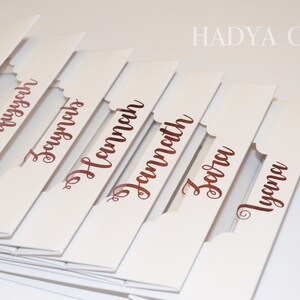 Puede incluir: Tarjetas de mesa blancas con bordes festoneados y nombres en escritura color cobre. Los nombres incluyen Amelie, Safiyyah, Zaynab, Hannah, Zara y Tyana. La marca, HADYA CO, est&aacute; impresa en la esquina superior derecha.