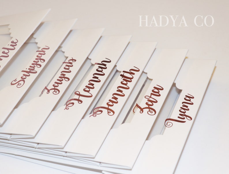 Puede incluir: Tarjetas de mesa blancas con bordes festoneados y nombres en escritura color cobre. Los nombres incluyen Amelie, Safiyyah, Zaynab, Hannah, Zara y Tyana. La marca, HADYA CO, est&aacute; impresa en la esquina superior derecha.