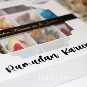 Puede incluir: Una caja blanca con tapa transparente, llena de caramelos de goma de colores. La caja est&aacute; dividida en compartimentos y tiene las palabras "Ramadan Kareem" en escritura negra. La marca "HADYA CO" est&aacute; impresa en la parte inferior.