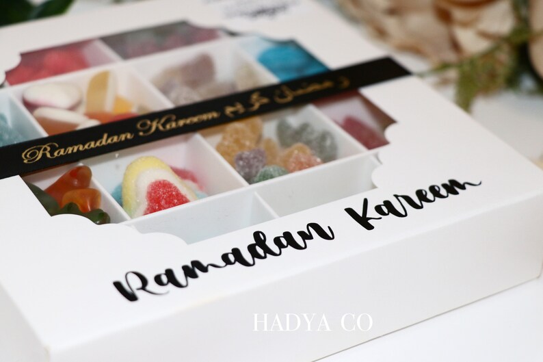 Puede incluir: Una caja blanca con tapa transparente, llena de caramelos de goma de colores. La caja est&aacute; dividida en compartimentos y tiene las palabras "Ramadan Kareem" en escritura negra. La marca "HADYA CO" est&aacute; impresa en la parte inferior.