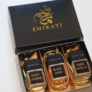 May include: A black box with gold lettering reading "EMIRATI", containing three perfume bottles. The bottles are labelled "Sweet Jasmine", "Black Oudh", and "Suede Vanilla". Each bottle has a gold frame and a gold cord.