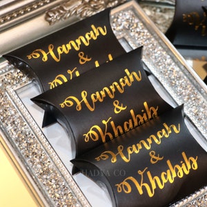 May include: Three black pillow boxes with gold lettering. The boxes are stacked on top of each other. The top box says "Hannah & ..." The middle box says "Hannah & Khabib". The bottom box says "Hannah & Khabib".
