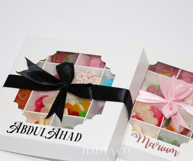 Puede incluir: Dos cajas de regalo blancas llenas de caramelos coloridos, cada una con una ventana transparente y una cinta decorativa. Una caja tiene una cinta negra y el nombre "ABDUL AHAD" impreso. La otra caja tiene una cinta rosa y el nombre "Mariam" impreso.