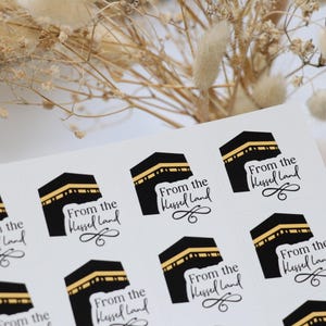 May include: A sheet of white stickers with black and gold Kaaba designs and the words "From the Blessed Land". The stickers are arranged on a white surface, with a blurred background.