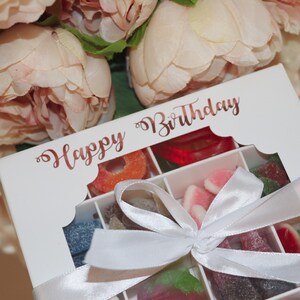Puede incluir: Una caja de regalo blanca llena de caramelos coloridos, atada con una cinta blanca. La caja tiene una ventana transparente y las palabras "Happy Birthday" en escritura oro rosa. Flores rosas suaves en el fondo.