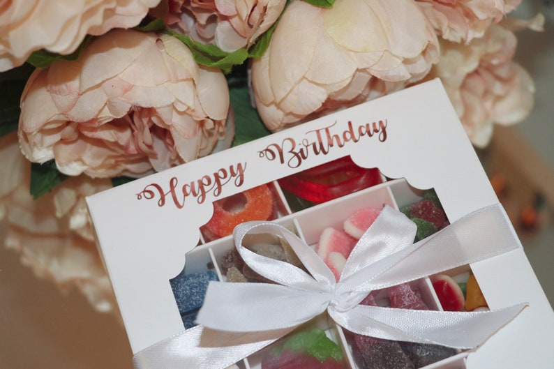 Puede incluir: Una caja de regalo blanca llena de caramelos coloridos, atada con una cinta blanca. La caja tiene una ventana transparente y las palabras "Happy Birthday" en escritura oro rosa. Flores rosas suaves en el fondo.