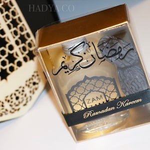 May include: A gold gift box with a clear lid containing a small bottle of perfume and a date. The bottle has a black label with Arabic text and the words "Zam Ramadan Kareem".