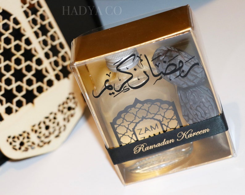 May include: A gold gift box with a clear lid containing a small bottle of perfume and a date. The bottle has a black label with Arabic text and the words "Zam Ramadan Kareem".